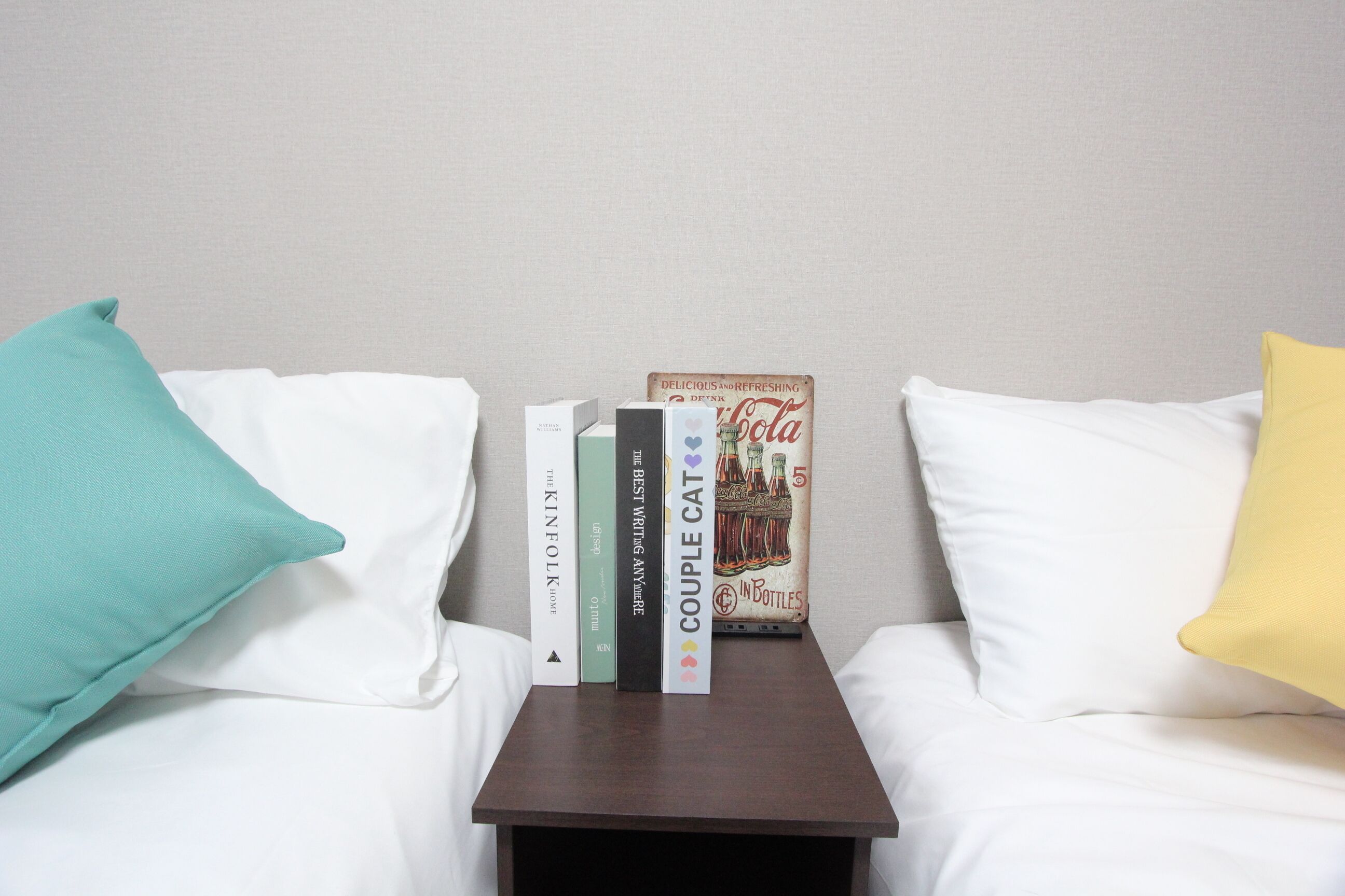 A neatly made bed with white sheets, a teal pillow, a yellow pillow, and a small wooden nightstand with books and a Coca-Cola poster.