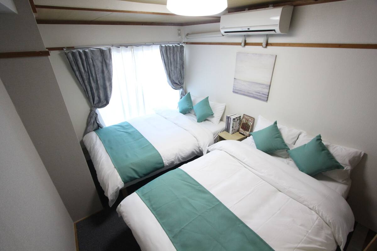 A hotel room with two beds, each with white and teal bedding, a nightstand with a lamp, and a window with curtains.