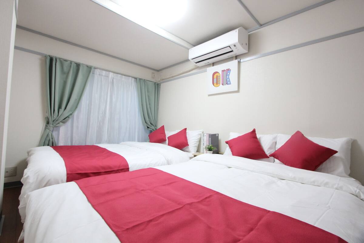 A hotel room with two beds, each with red and white bedding, an air conditioning unit, and a window with curtains.