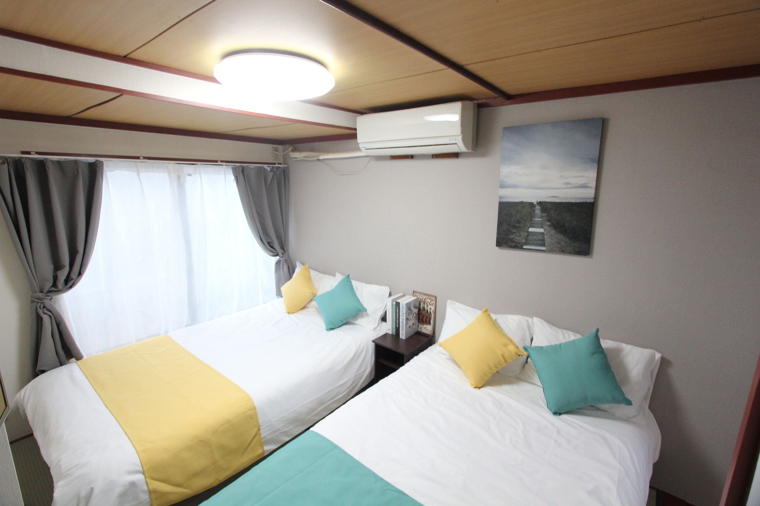 Une chambre d’hôtel avec deux lits, chacun agrémenté de draps blancs et d’oreillers jaunes et turquoise. On y trouve un luminaire au plafond, un appareil de climatisation et un tableau encadré accroché au mur.
