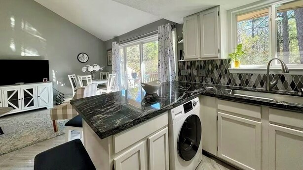 Cocina moderna con lavadora integrada, armarios blancos, encimera de mármol y una ventana con cortinas.