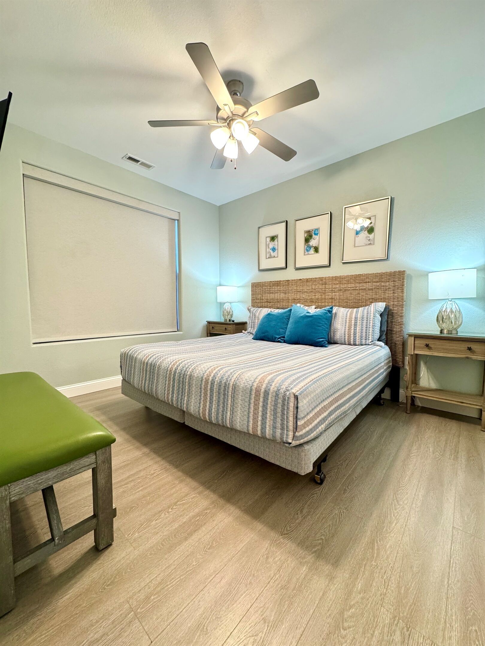 A bedroom with a bed, a green bench, a ceiling fan, and framed pictures on the wall.