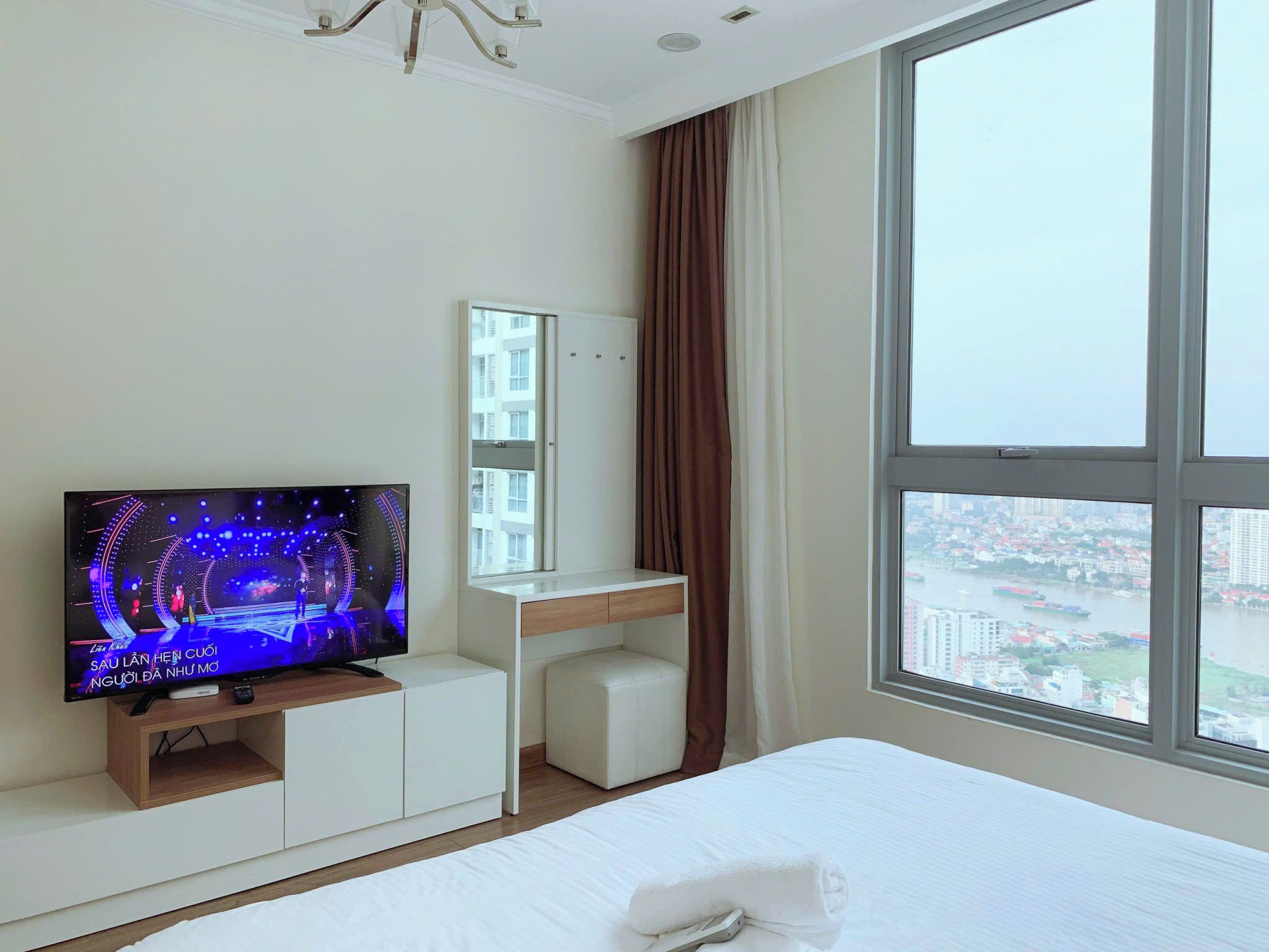A hotel room with a large bed, a TV on a wooden stand, a white desk, a mirror, and a window with a city view.