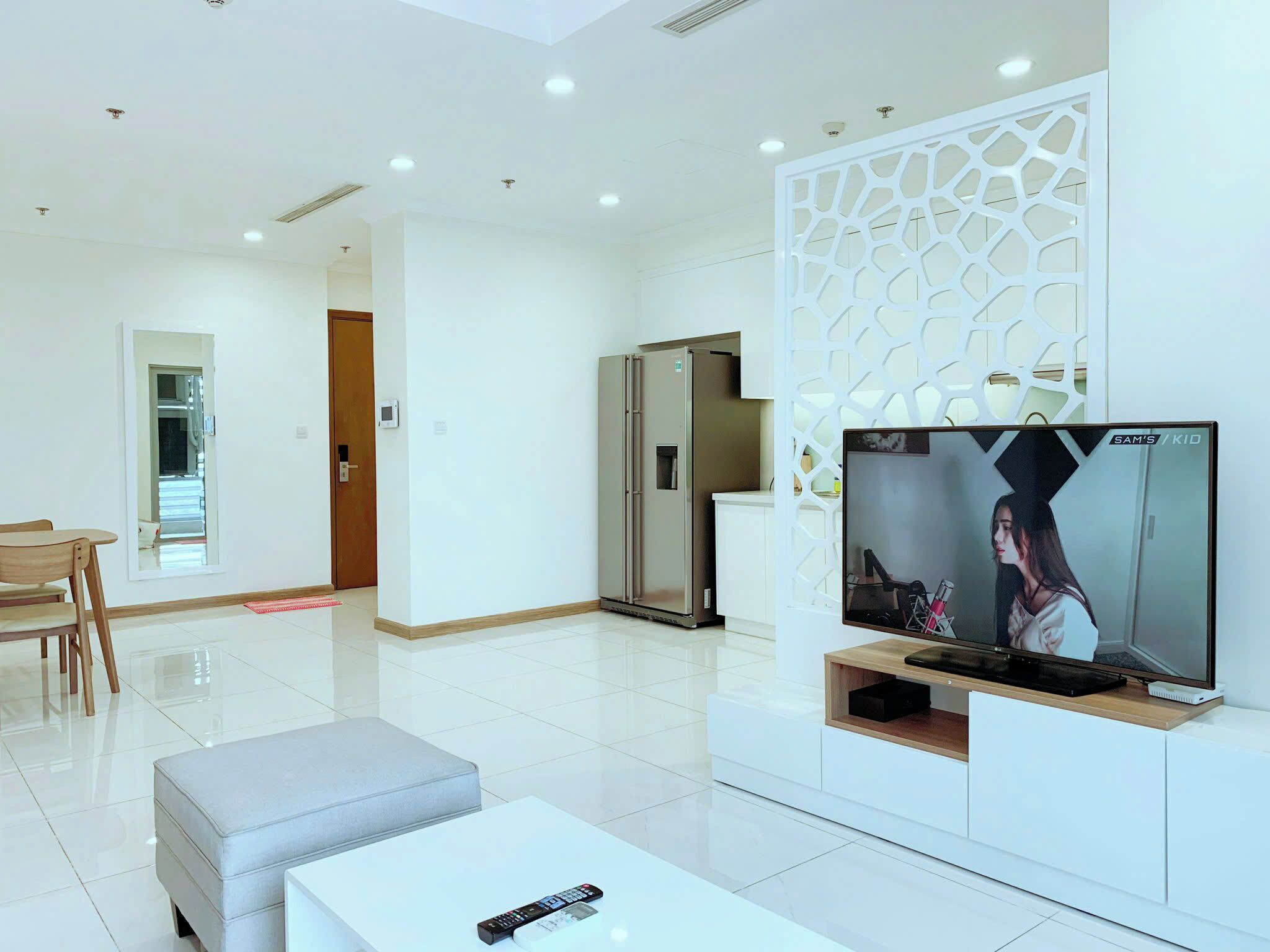 A modern living room with a flat-screen TV, a dining area with a table and chairs, and a kitchen area with a refrigerator.