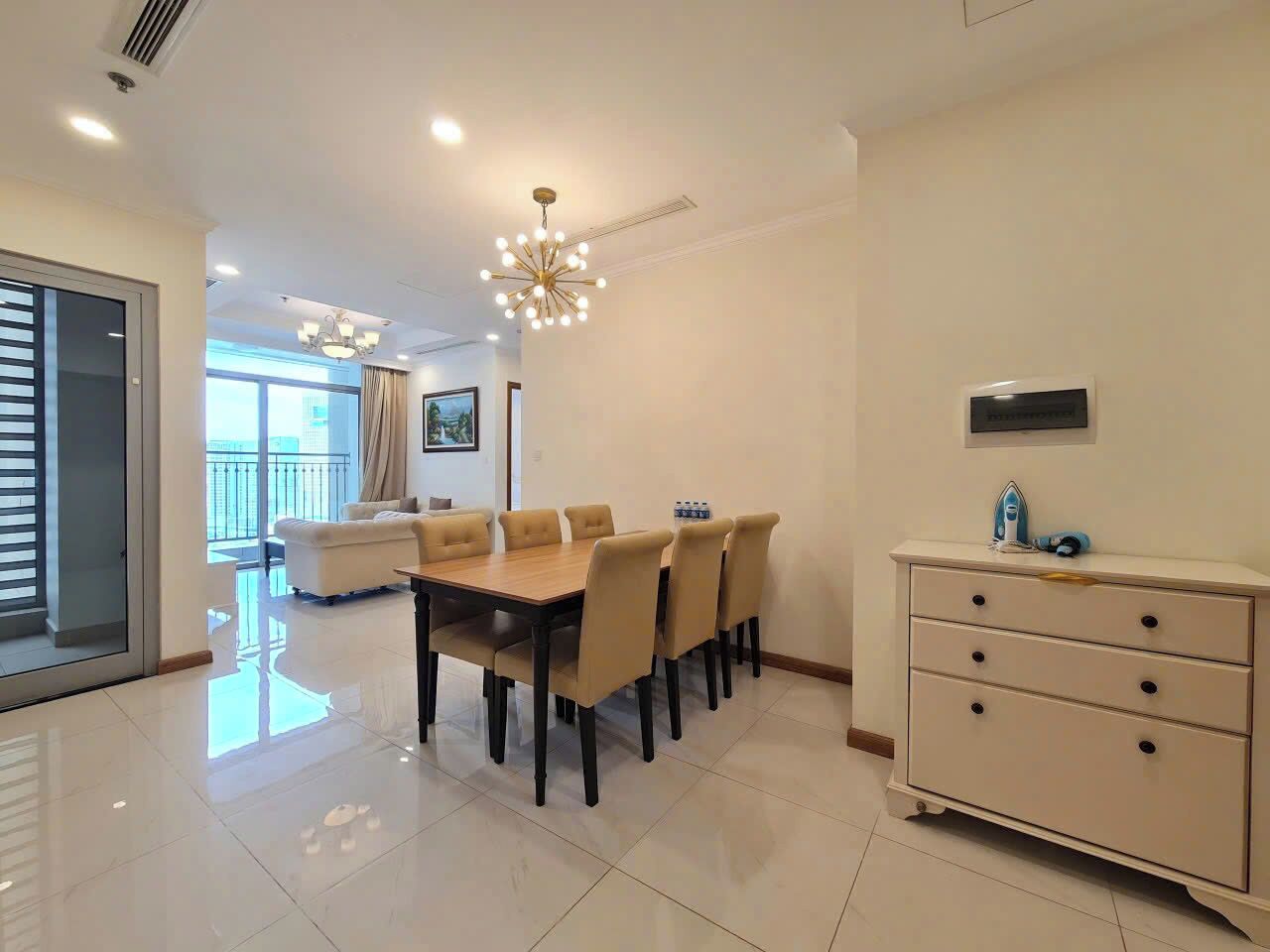 A modern dining area with a wooden table and beige chairs, a white dresser with drawers, and a chandelier.