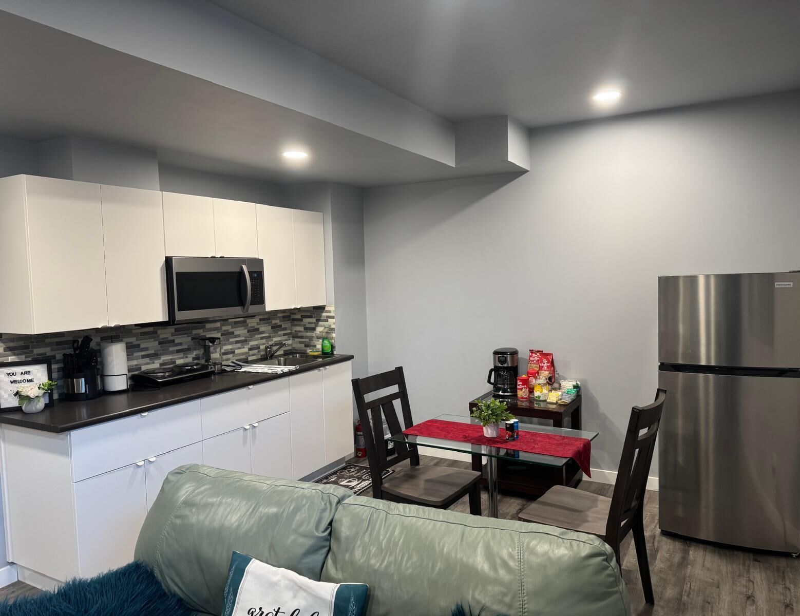 A modern kitchen with white cabinets, a black countertop, a stainless steel refrigerator, and a dining area with a glass table and chairs.