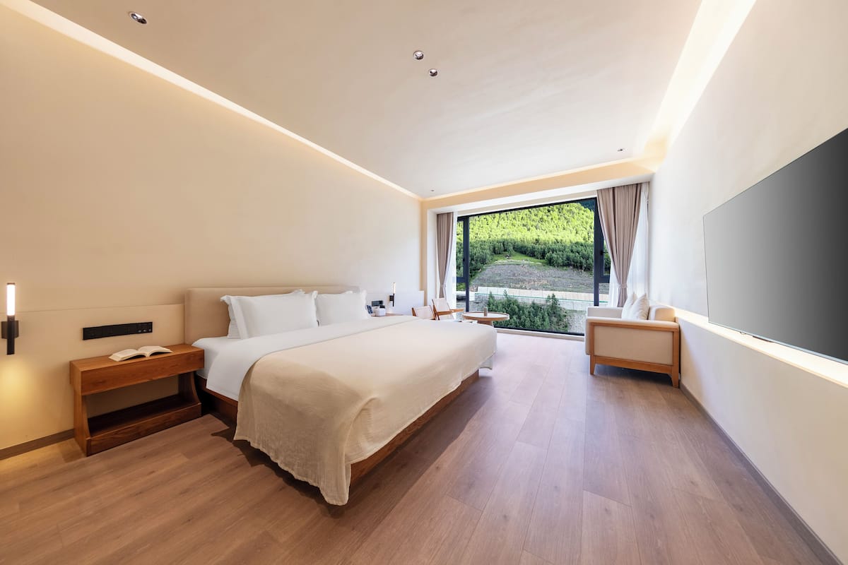 Ein modernes Hotelzimmer mit einem großen Bett, Nachttischen, einem Fernseher und Blick auf Grün hinter großen Fenstern.
