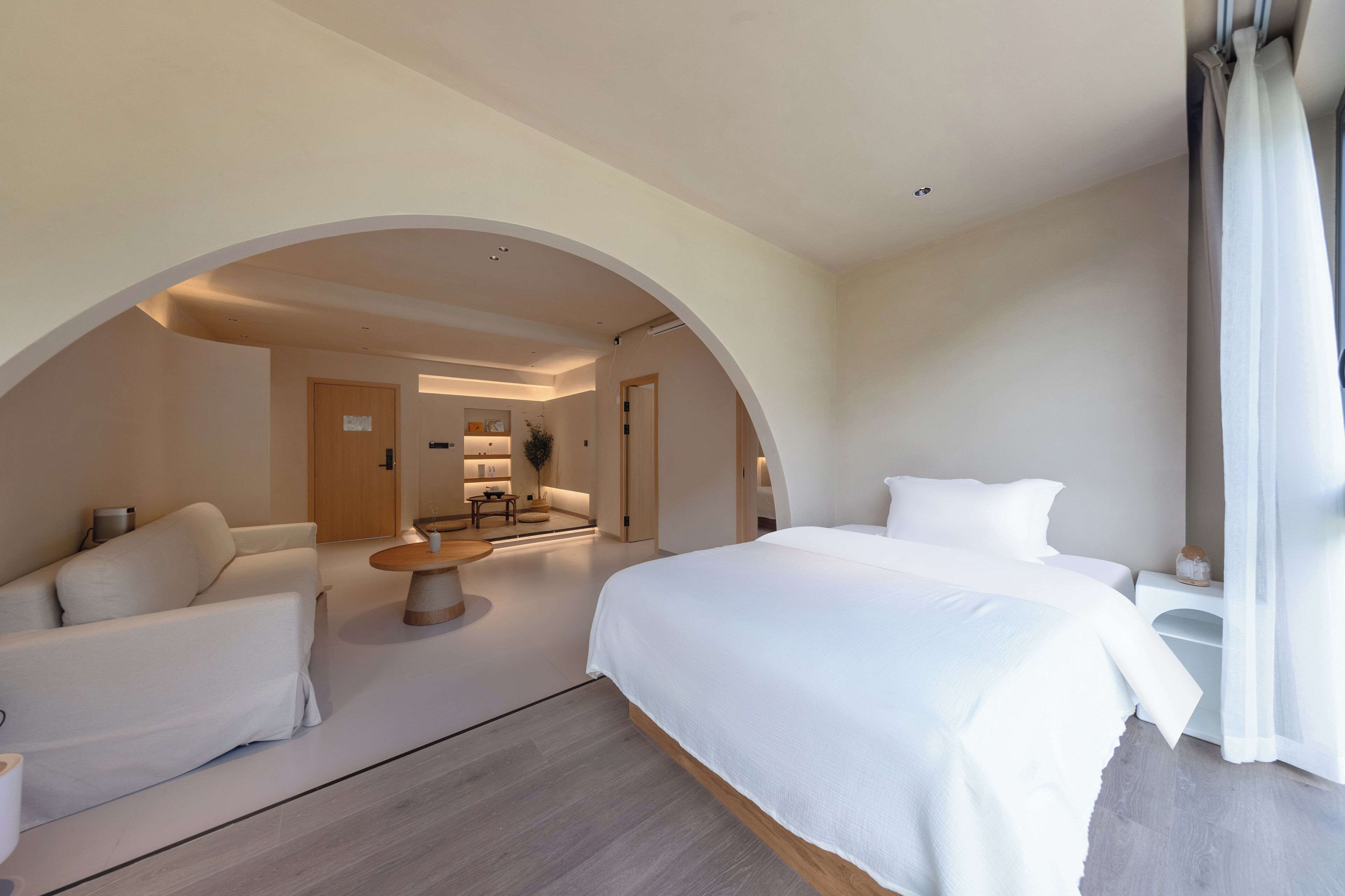 Quarto de hotel moderno com uma cama grande, um sofá, uma pequena mesa e vista para um banheiro através de uma porta em arco.