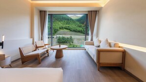 A modern hotel room with a large window offering a scenic mountain view. The room includes a sofa, two chairs, a round table, and a bed.