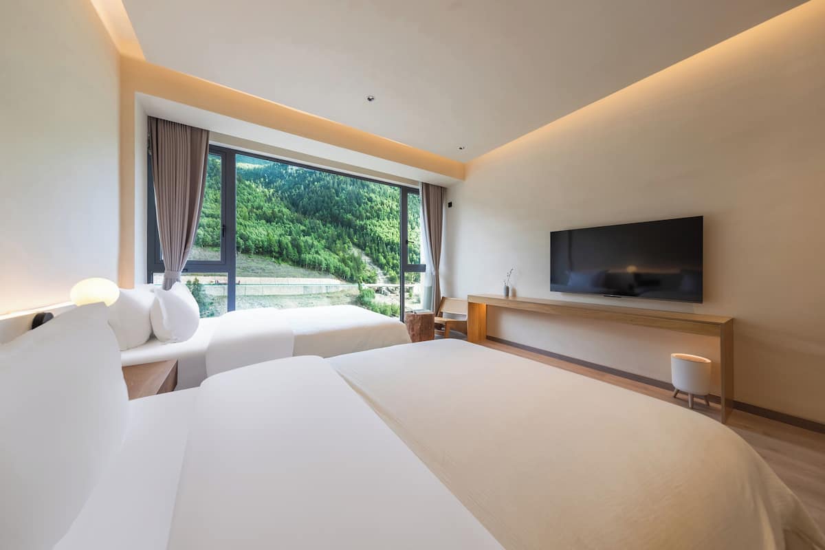 Ein modernes Hotelzimmer mit zwei Betten, einem Flachbildfernseher, einem kleinen Tisch und einem großen Fenster mit Blick auf Grünflächen.