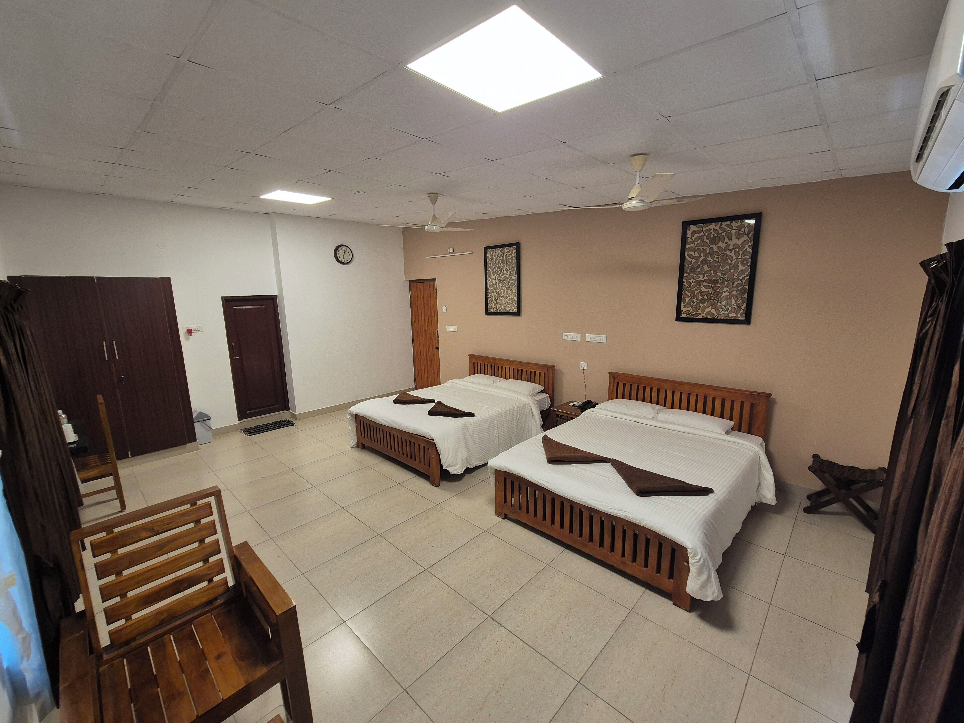A hotel room with two beds, a wooden chair, a wardrobe, a ceiling fan, and a clock on the wall.