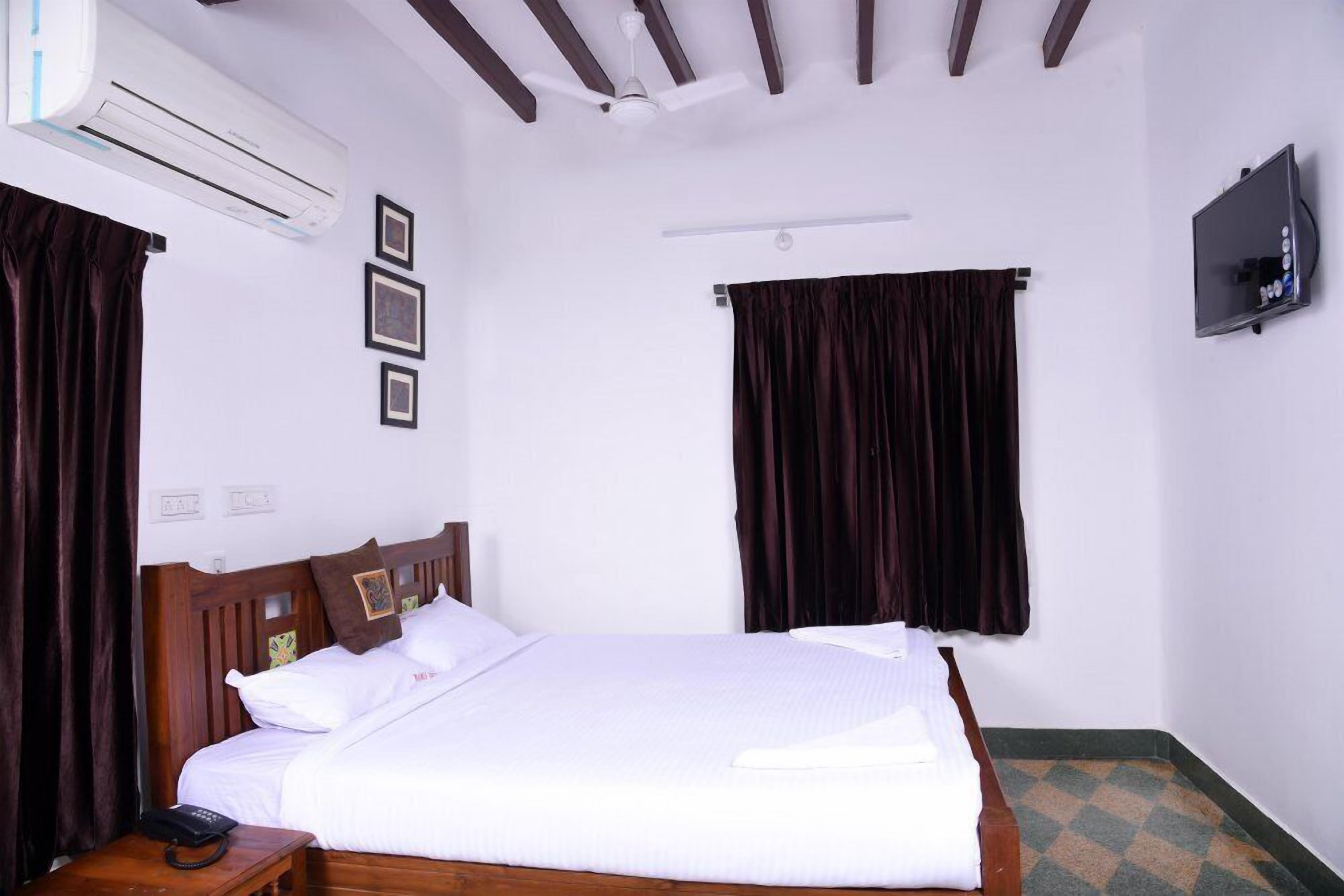 Une chambre d’hôtel avec un grand lit, un appareil de climatisation, un ventilateur de plafond, une télévision et un téléphone sur une table de chevet.