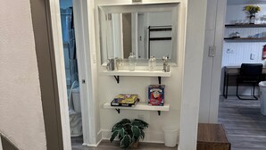 A hallway with a white wall, a mirror, a shelf with toiletries, a potted plant, and a trash bin. - 15 mins to Notre Dame; visit The Cozy Blue Haven in Niles, MI (Niles)