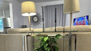A modern living room with a sofa, two side tables with lamps, a large potted plant, and a television displaying a game show. - 15 mins to Notre Dame; visit The Cozy Blue Haven in Niles, MI (Niles)