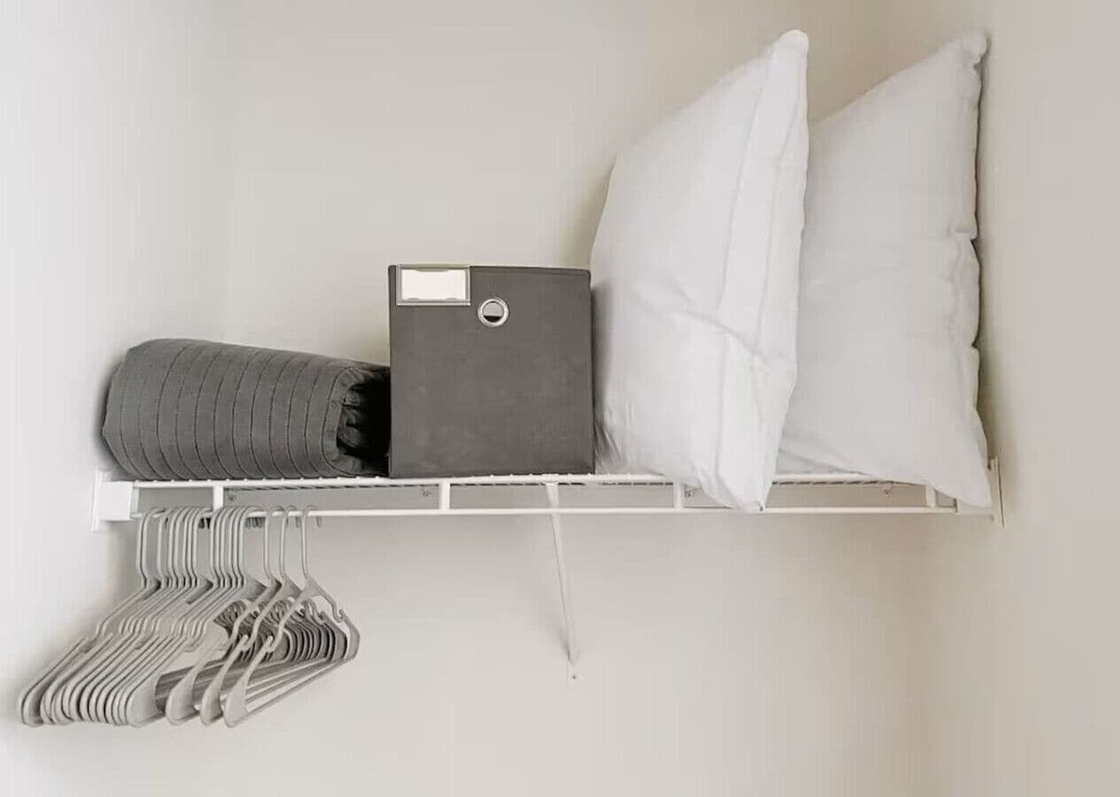 A white wall with a shelf holding a grey storage box, a rolled-up grey blanket, a white pillow, and a set of white and grey hangers.