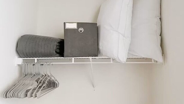 A white wall with a shelf holding a grey storage box, a rolled-up grey blanket, a white pillow, and a set of white and grey hangers.