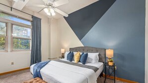 Ein Schlafzimmer mit einem Bett, zwei Nachttischen, einer Bank, einem Deckenventilator und einem Fenster mit Vorhängen.