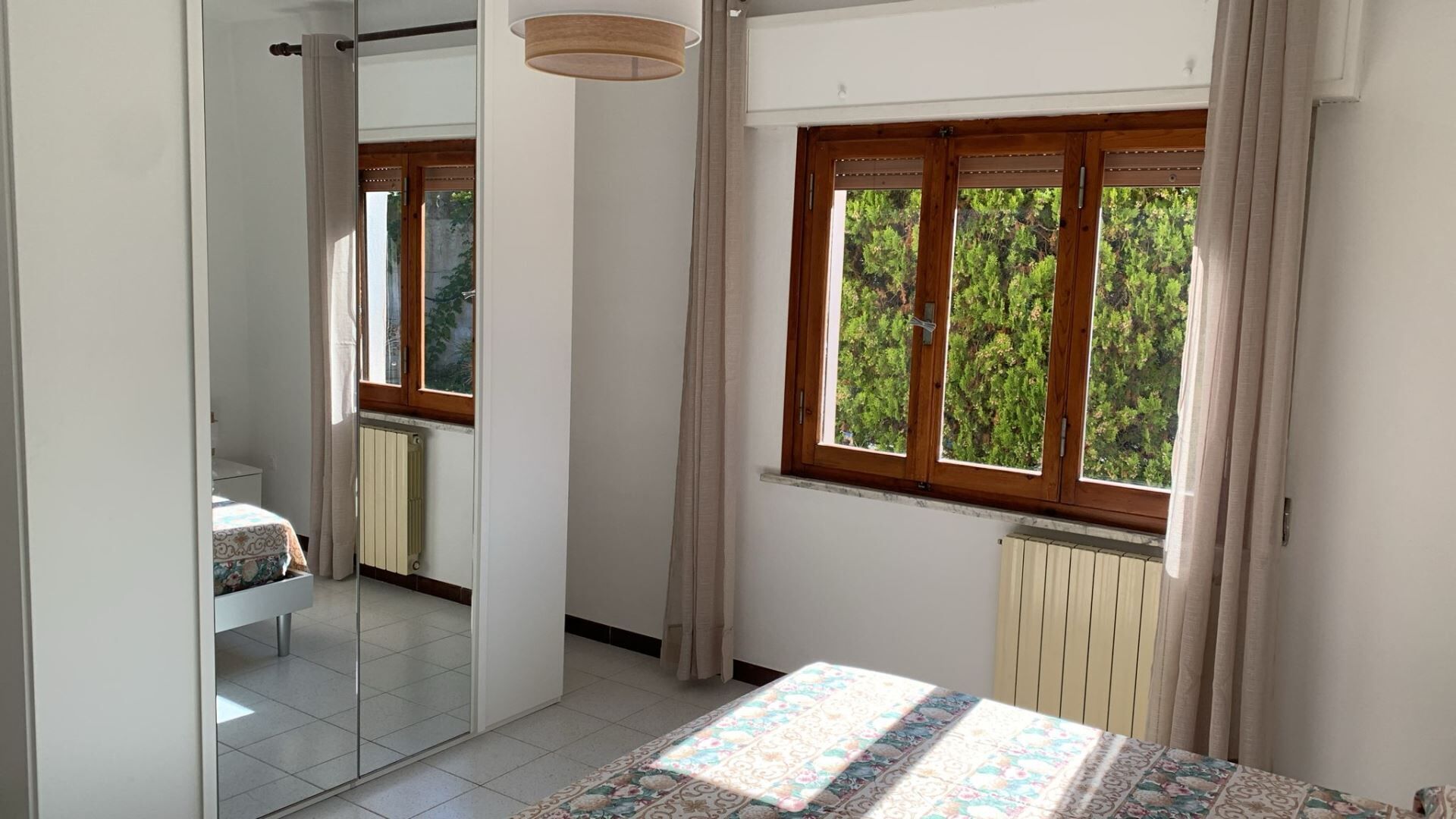 Una camera da letto con un letto, una finestra con tende, un termosifone e uno specchio.