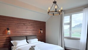 Een slaapkamer met een houten hoofdeinde, een groot bed met witte lakens, twee wandlampen en uitzicht op een landschap door een raam.