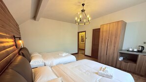 A hotel room with two beds, a wooden headboard, a chandelier, a wardrobe, and a small kitchenette.