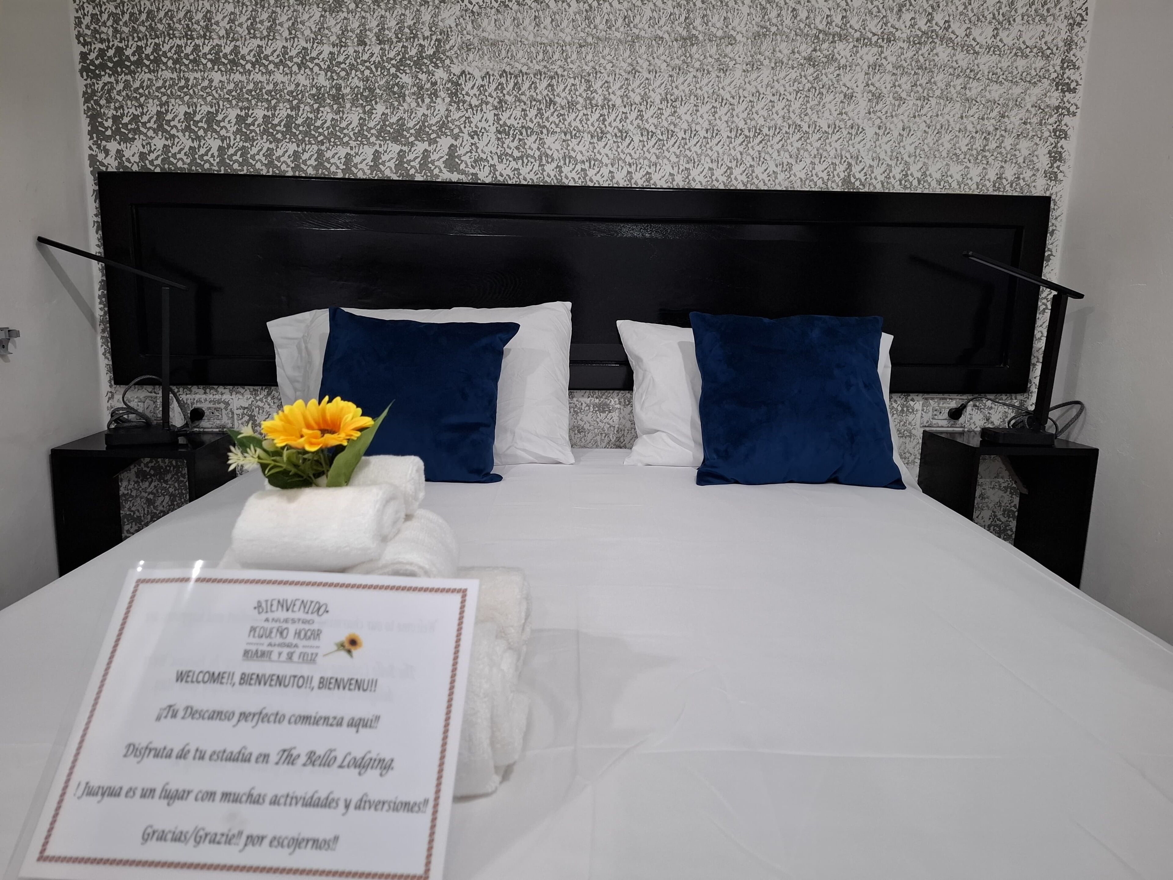 Una cama bien hecha con almohadas blancas y azules, una toalla doblada y una tarjeta de bienvenida.