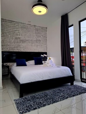 A modern hotel room with a large bed, a headboard with a textured design, a black nightstand, a white bedside table, and a black and white patterned rug. - The Bello Lodging A-Luxurious Studio Apartment (Juayua)