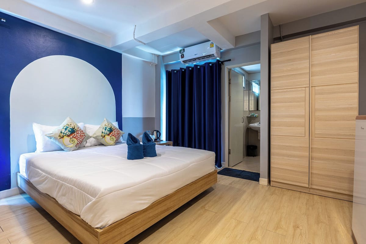 Une chambre moderne avec un grand lit, un ventilateur de plafond, une armoire intégrée et une salle de bain visible à travers une porte en verre.