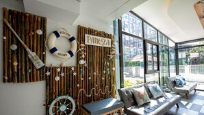 A room with a nautical theme, featuring a bamboo wall with a 'Welcome Aboard' sign, a life ring, and a ship's wheel. There are two grey sofas with cushions and a bench. - PanoSea Bangsaen (Chonburi)