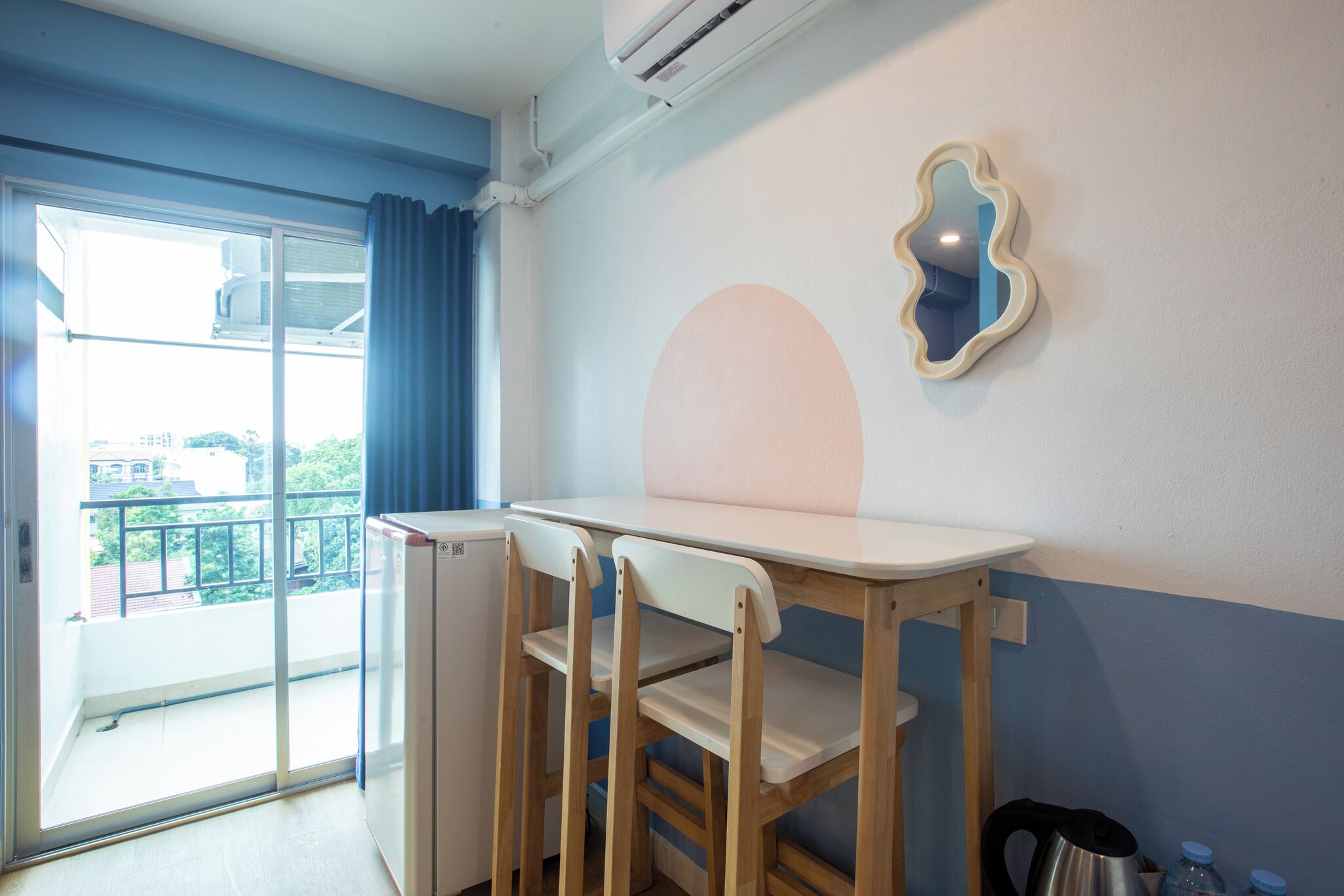 A room with a wooden table and chairs, a small refrigerator, a wall-mounted mirror, and a view of a balcony.