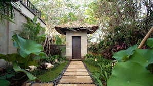 A pathway leading to a small building with a thatched roof, surrounded by lush greenery and large leaves. - Sundara Oceanside Villa (Tabanan)