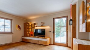 A spacious living room with a large TV, wooden cabinets, and a view of trees through the sliding glass door. - Detached holiday home with terrace and balcony. (Bramberg am Wildkogel)