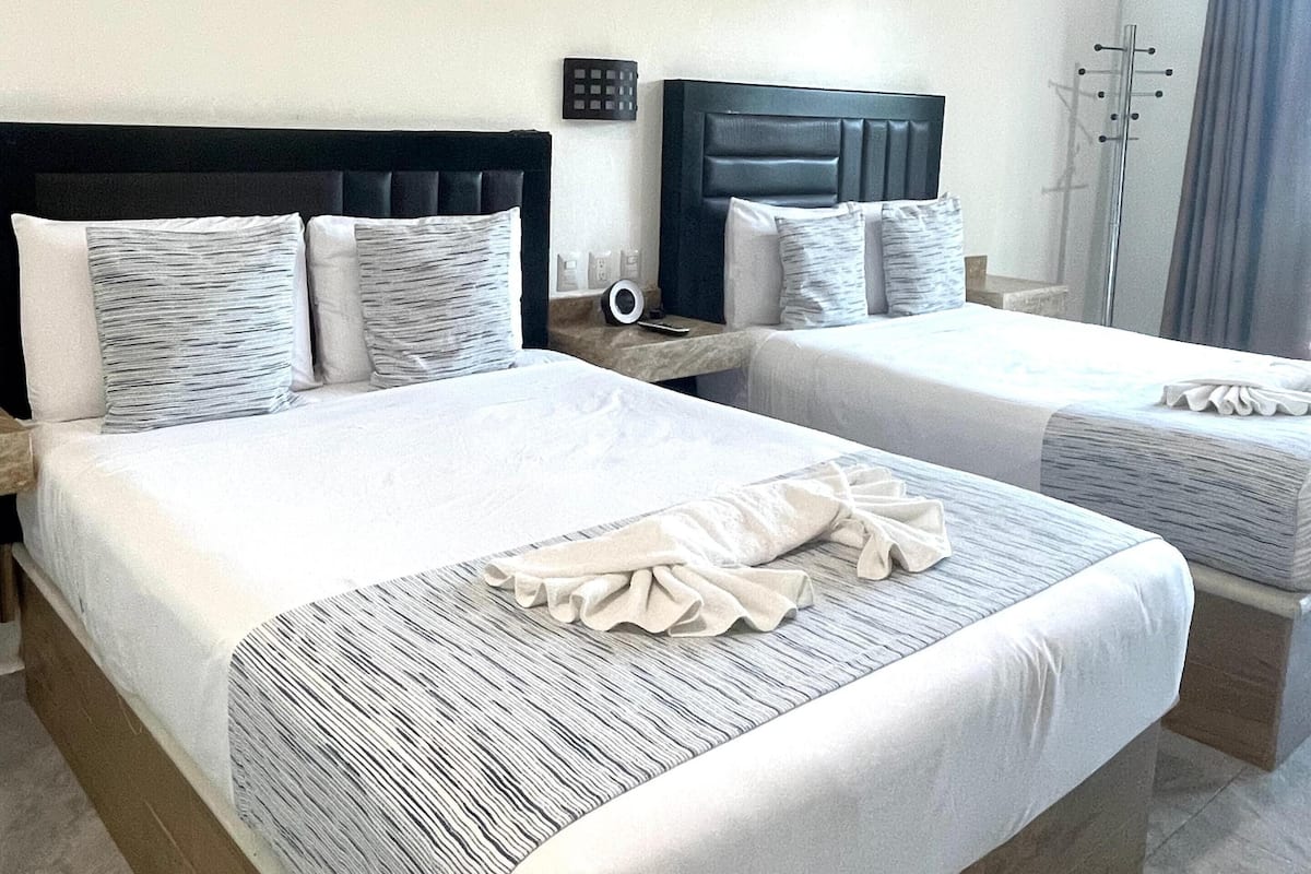 A hotel room with two beds, a dark headboard, a bedside table with a lamp, and an air conditioning unit.