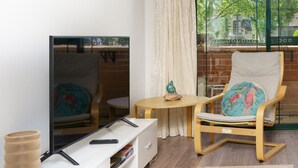 Un salon moderne avec un téléviseur à écran plat, une chaise en bois avec un coussin coloré, une petite table ronde et un meuble blanc contenant des livres.