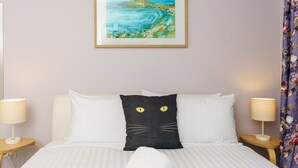 Une chambre d’hôtel avec un lit, des oreillers blancs, un coussin représentant un chat noir, deux lampes de chevet, un tableau encadré et un rideau à motifs floraux.