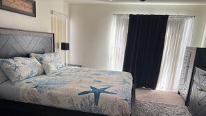 Quarto com cama, mesa de cabeceira, cômoda e janela com cortinas.