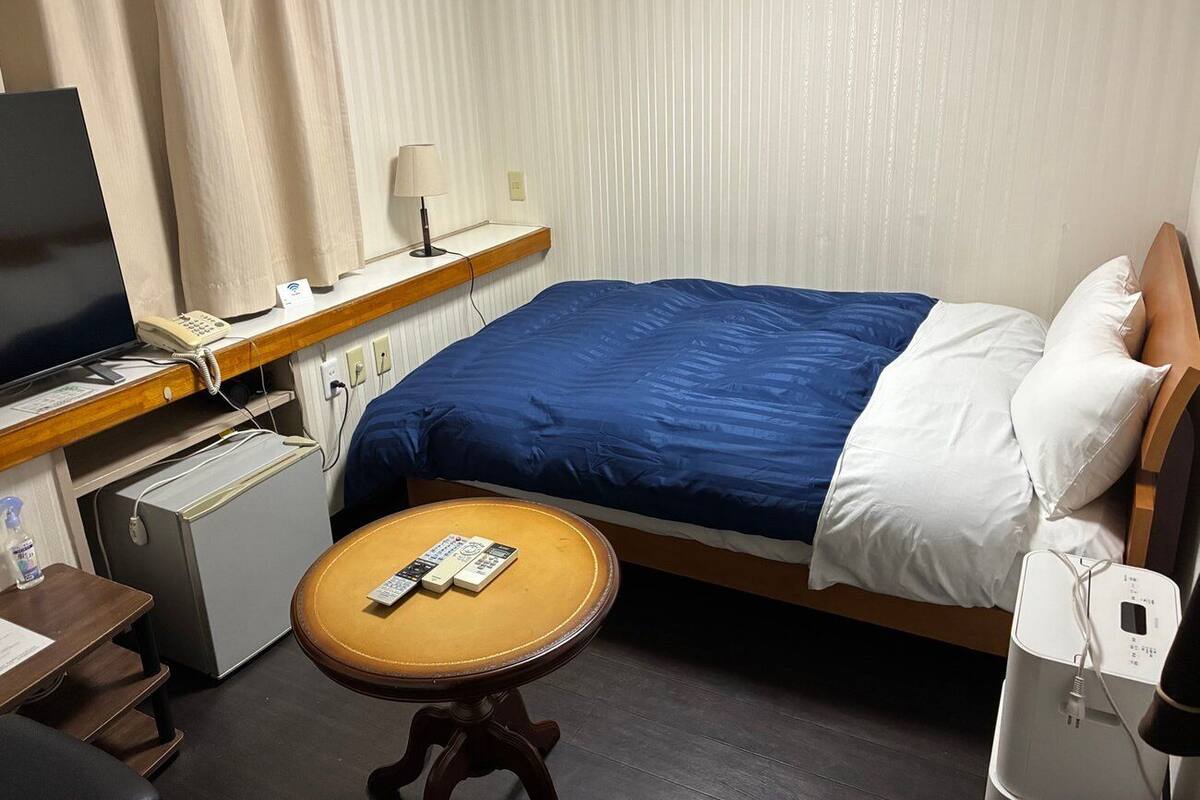 Hotelkamer met een bed, een bureau met een computer, een kleine koelkast, een nachtkastje met een afstandsbediening en een lamp.