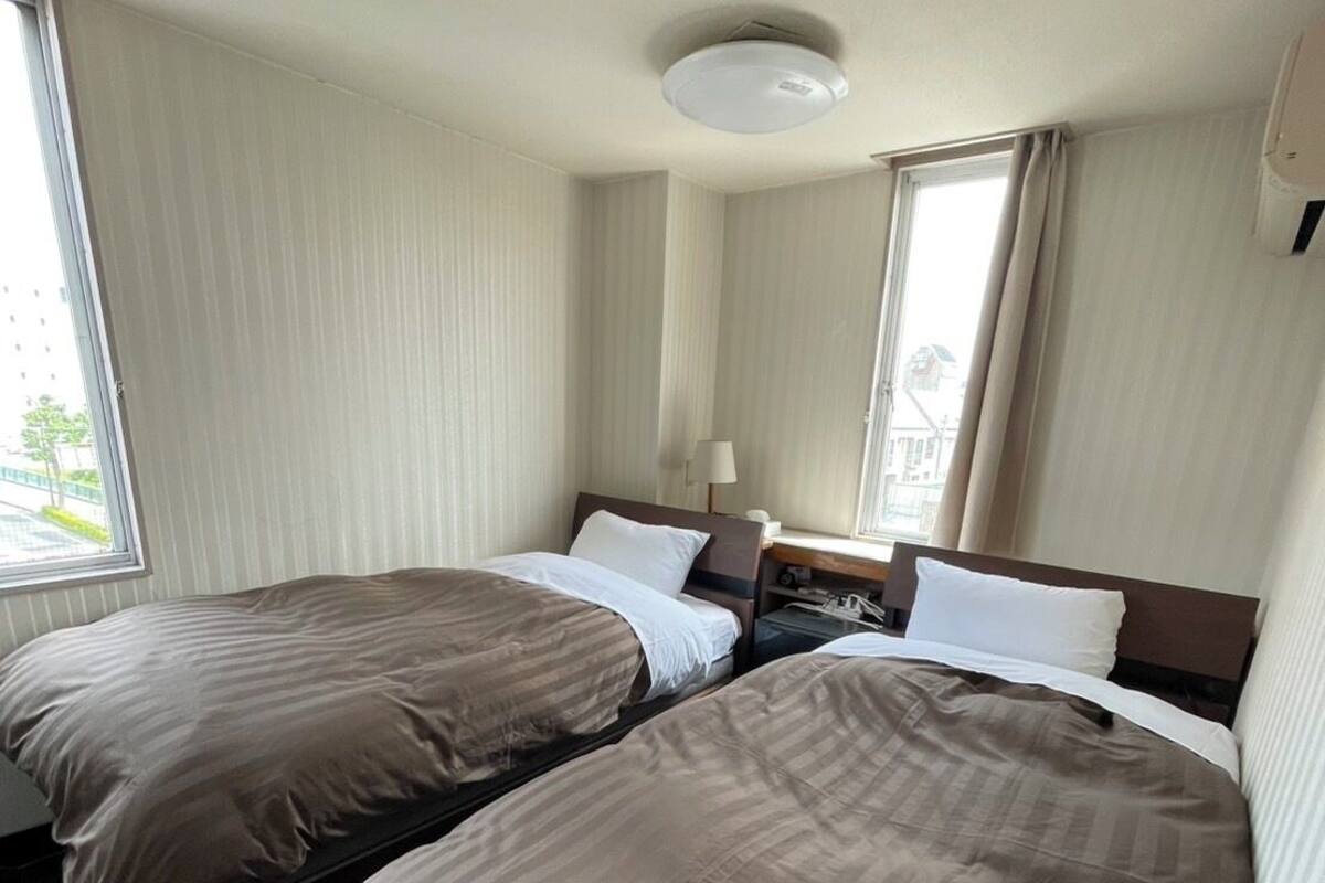 Een hotelkamer met twee enkele bedden, een bureau, een stoel, een lamp en een raam met gordijnen.