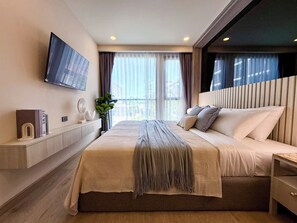 A modern hotel room with a large bed, a flat-screen TV mounted on the wall, a bedside table, and a window with curtains. - The Ozone Rezidence (Choeng Thale)