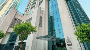 A modern high-rise building with a glass facade, a central entrance, and a landscaped area with trees. - Act 1 Act 2 Towers by DVH Luxe Retreats (Dubai)