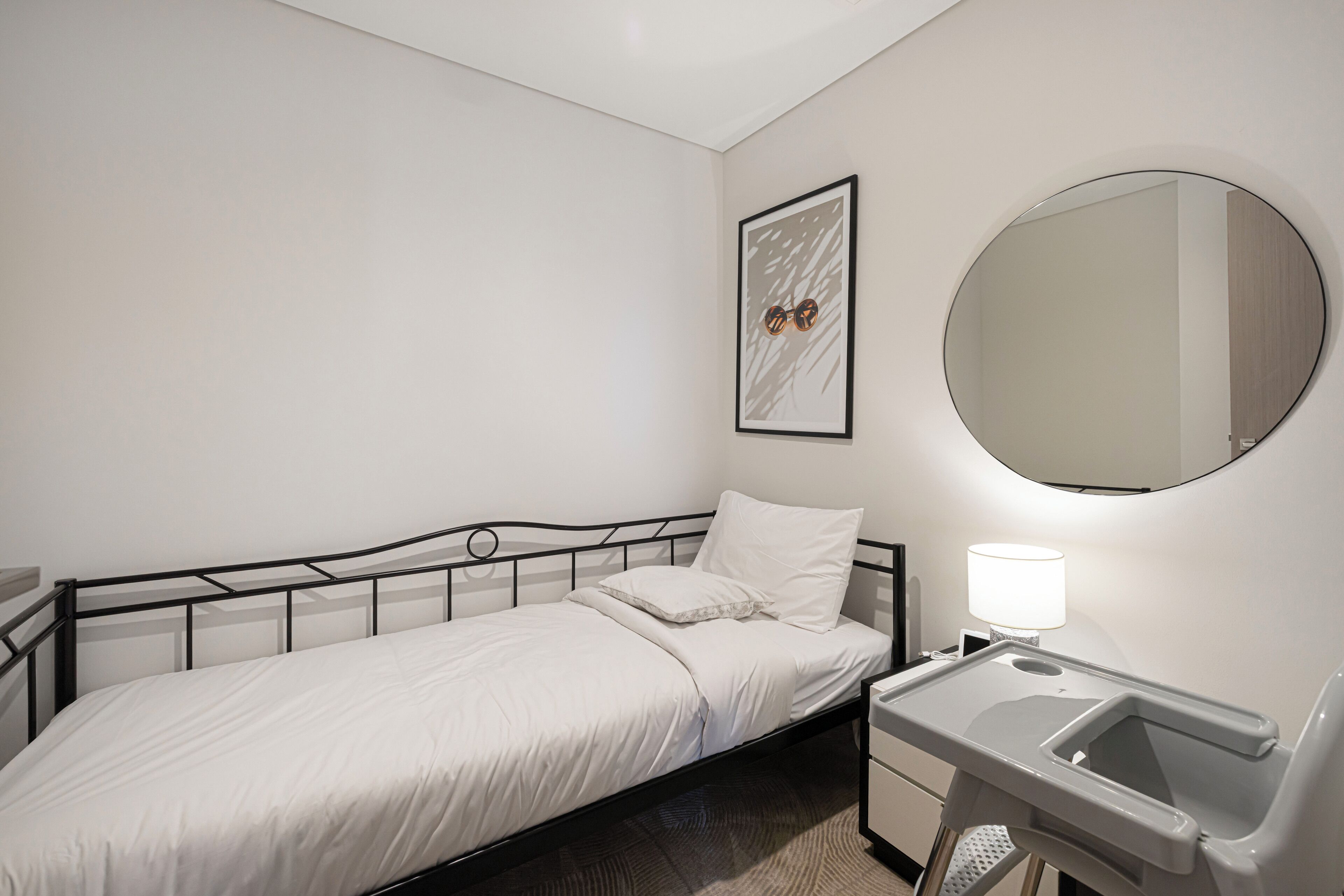 A single bed with a headboard, a bedside table with a lamp, a round mirror, and a framed picture on the wall.