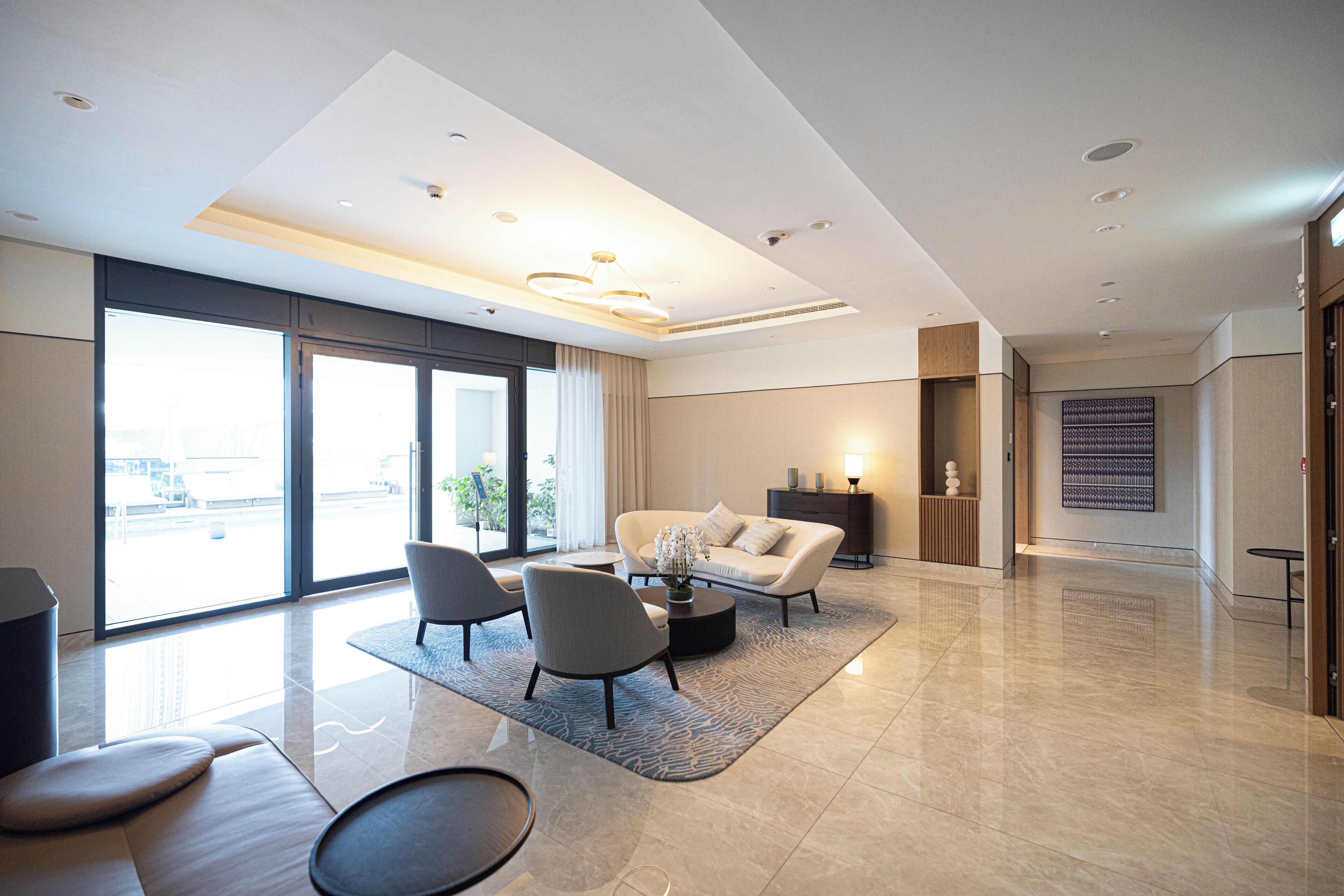 A modern living room with a sofa, chairs, a coffee table, and a rug. There is a large window with curtains, a lamp, and a wall-mounted artwork.