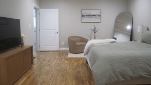 A bedroom with a wooden floor, a bed with a grey blanket, a TV on a wooden cabinet, a beige armchair, a white door, a painting on the wall, and a mirror.