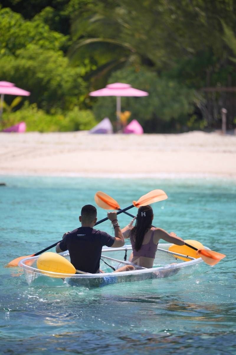 Two people in a kayak on clear water with a beach and umbrellas in the background.