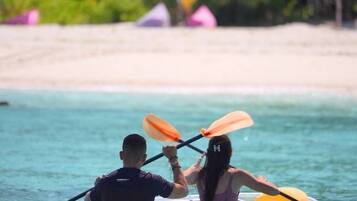 Two people in a kayak on clear water with a beach and umbrellas in the background.