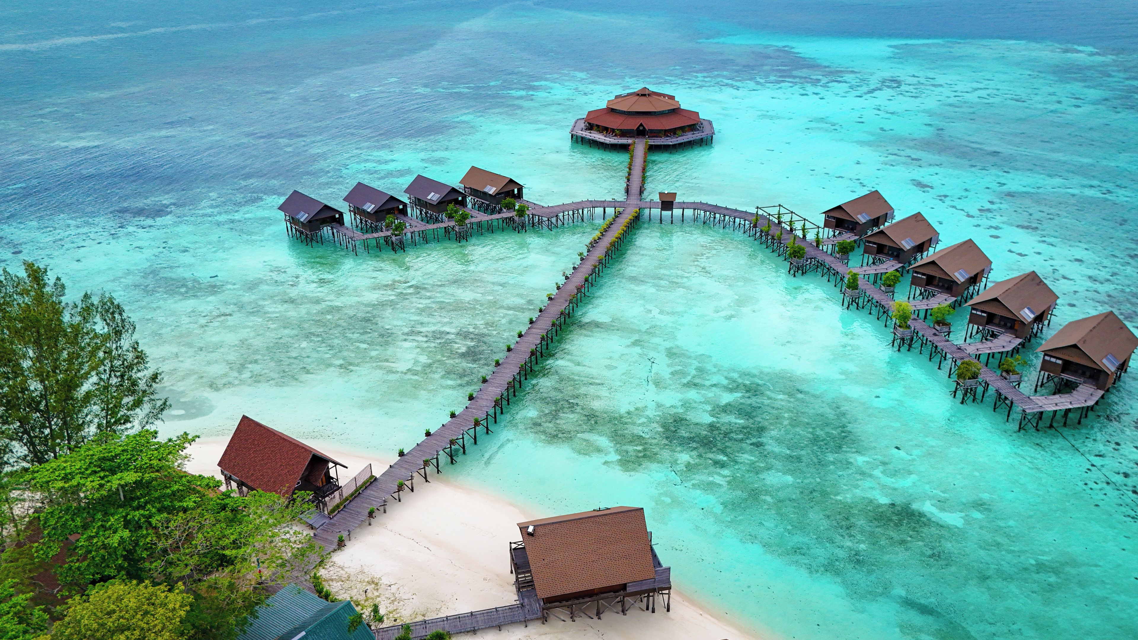 An aerial view of a resort with overwater bungalows connected by a long pier, clear turquoise waters, and a sandy beach.