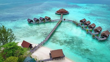 An aerial view of a resort with overwater bungalows connected by a long pier, clear turquoise waters, and a sandy beach.