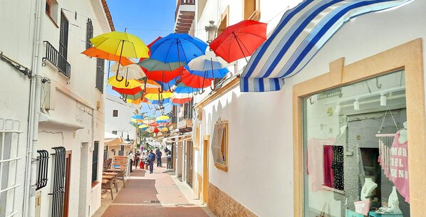 Une rue étroite, au-dessus de laquelle sont suspendus des parasols colorés, avec un magasin sur la droite et, sur la gauche, une enseigne indiquant les locations de vacances de SOLIDAYS.es.