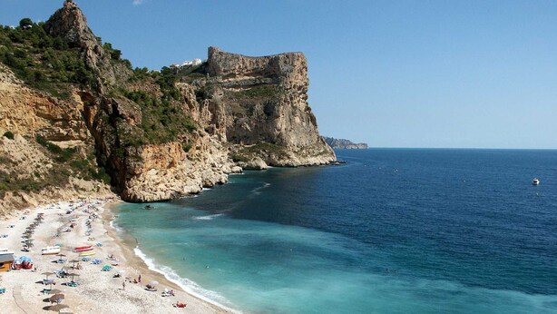 Une plage côtière aux eaux turquoise et limpides, bordée d’une falaise rocheuse et surmontée d’une structure qui ressemble à un château.