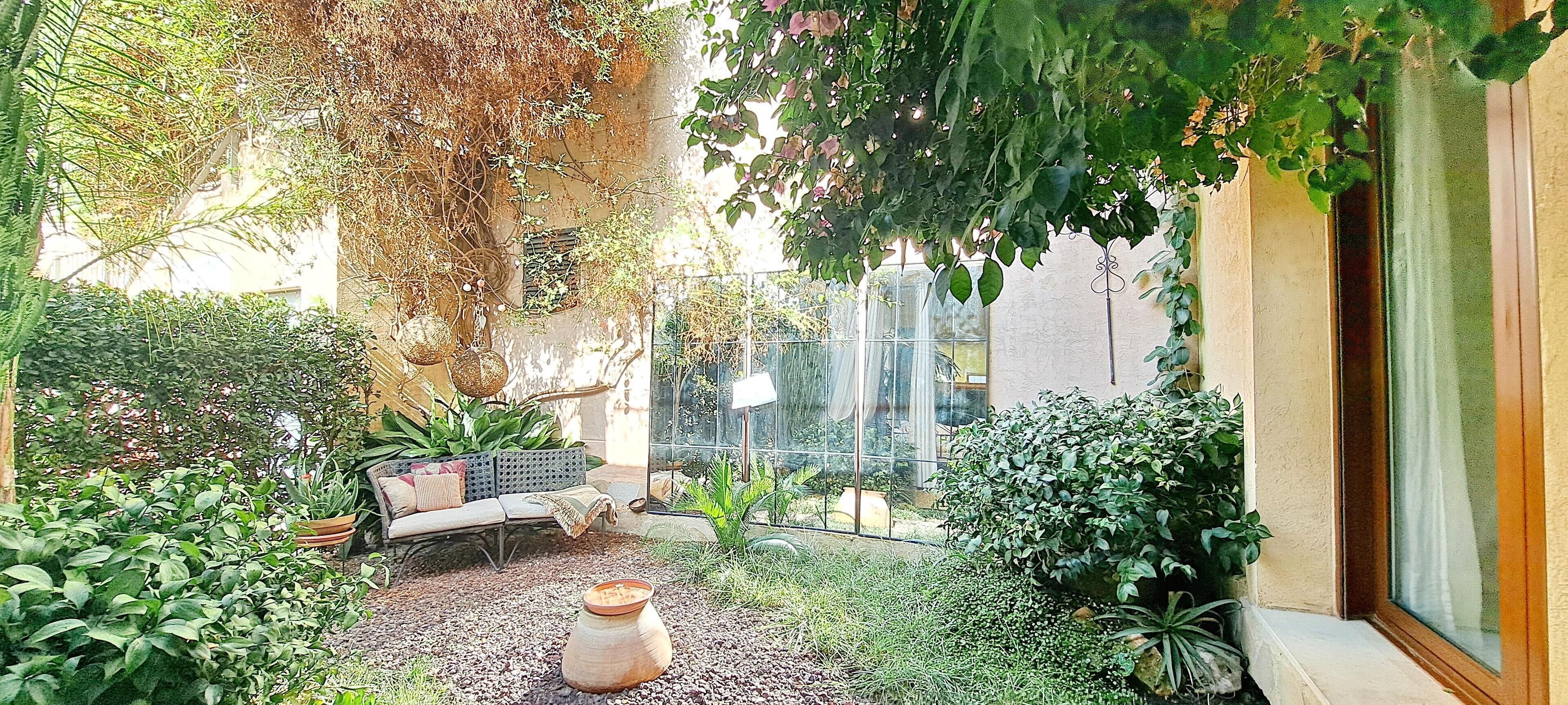 Un espace extérieur accueillant, aménagé avec un canapé et une table, entouré d’une végétation luxuriante et agrémenté d’un pot décoratif.