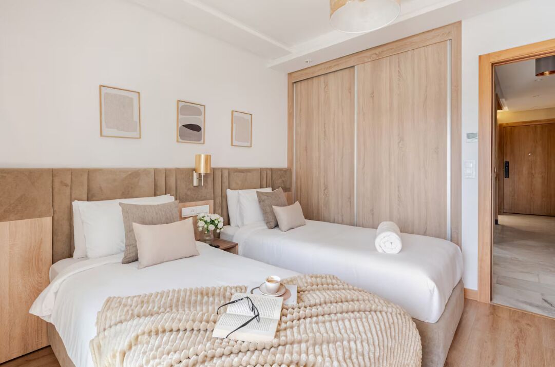 A hotel room with two beds, a wooden wardrobe, a bedside table with a lamp, and framed pictures on the wall.